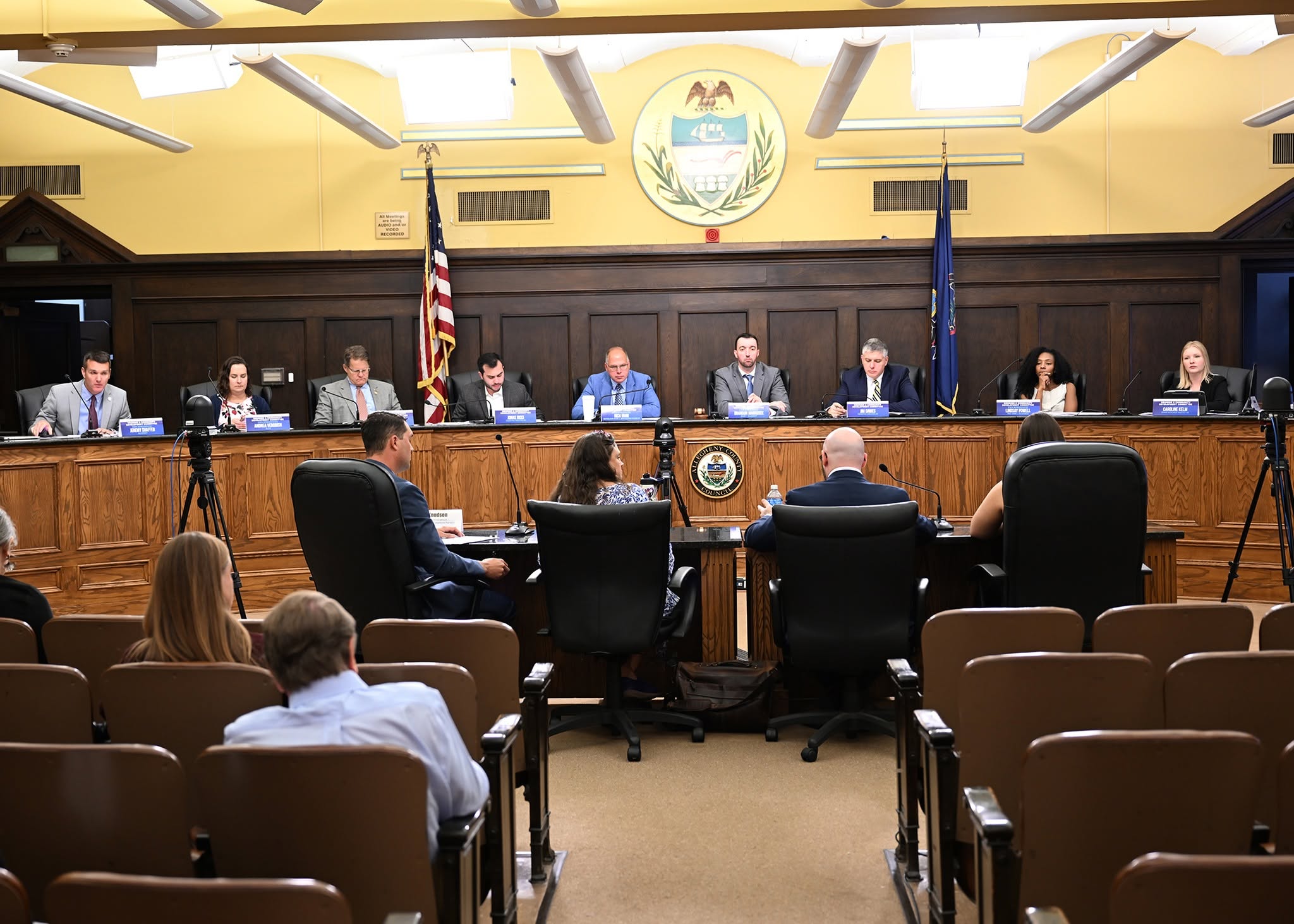A photo of a hearing with elected officials at a rostrum seated facing a table with four panelists addressing them.